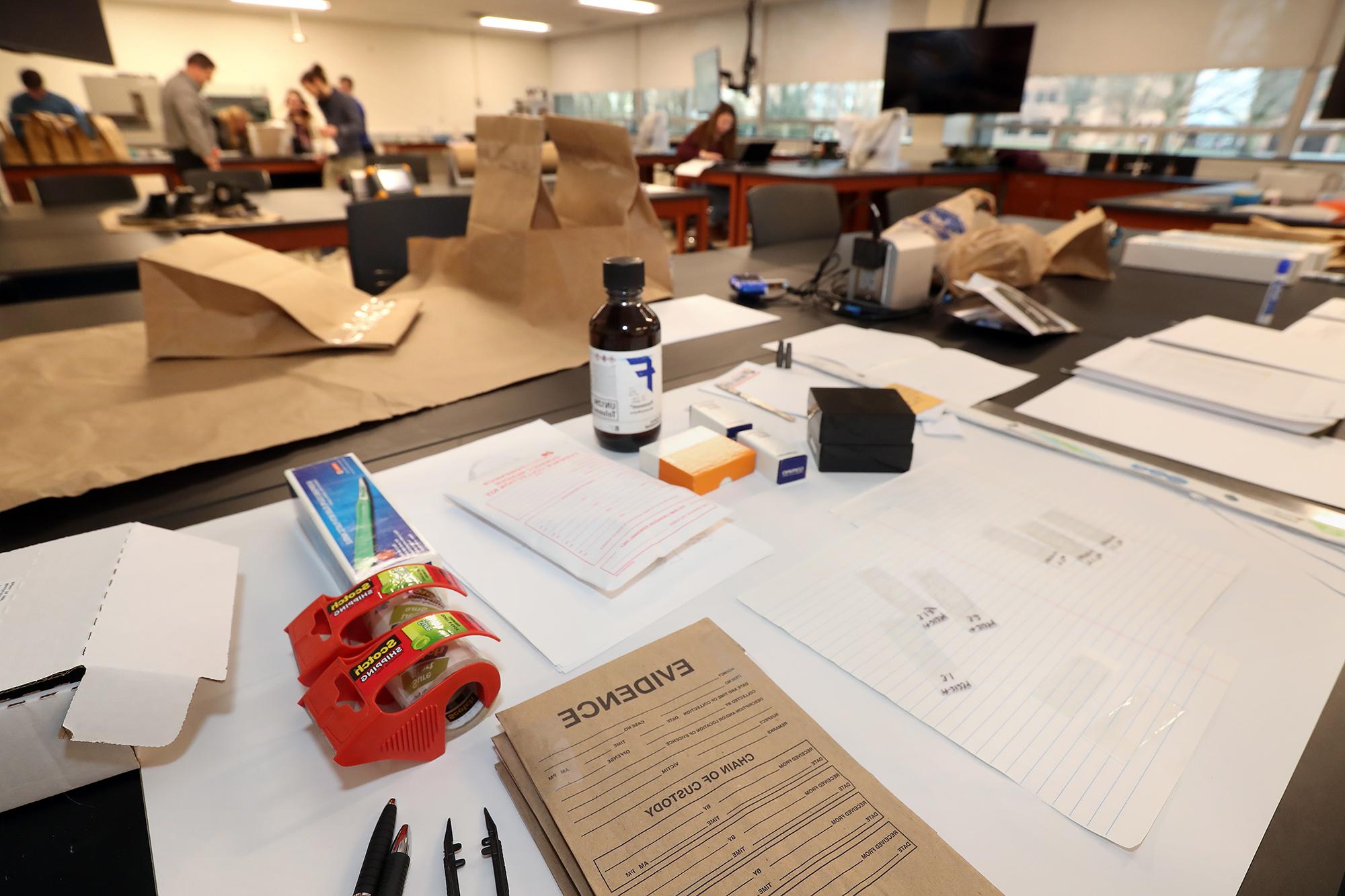 A table 在 BGSU forensic lab features brown paper evidence bags, chemicals and others tools that the students 在 background use to process crime scene evidence.