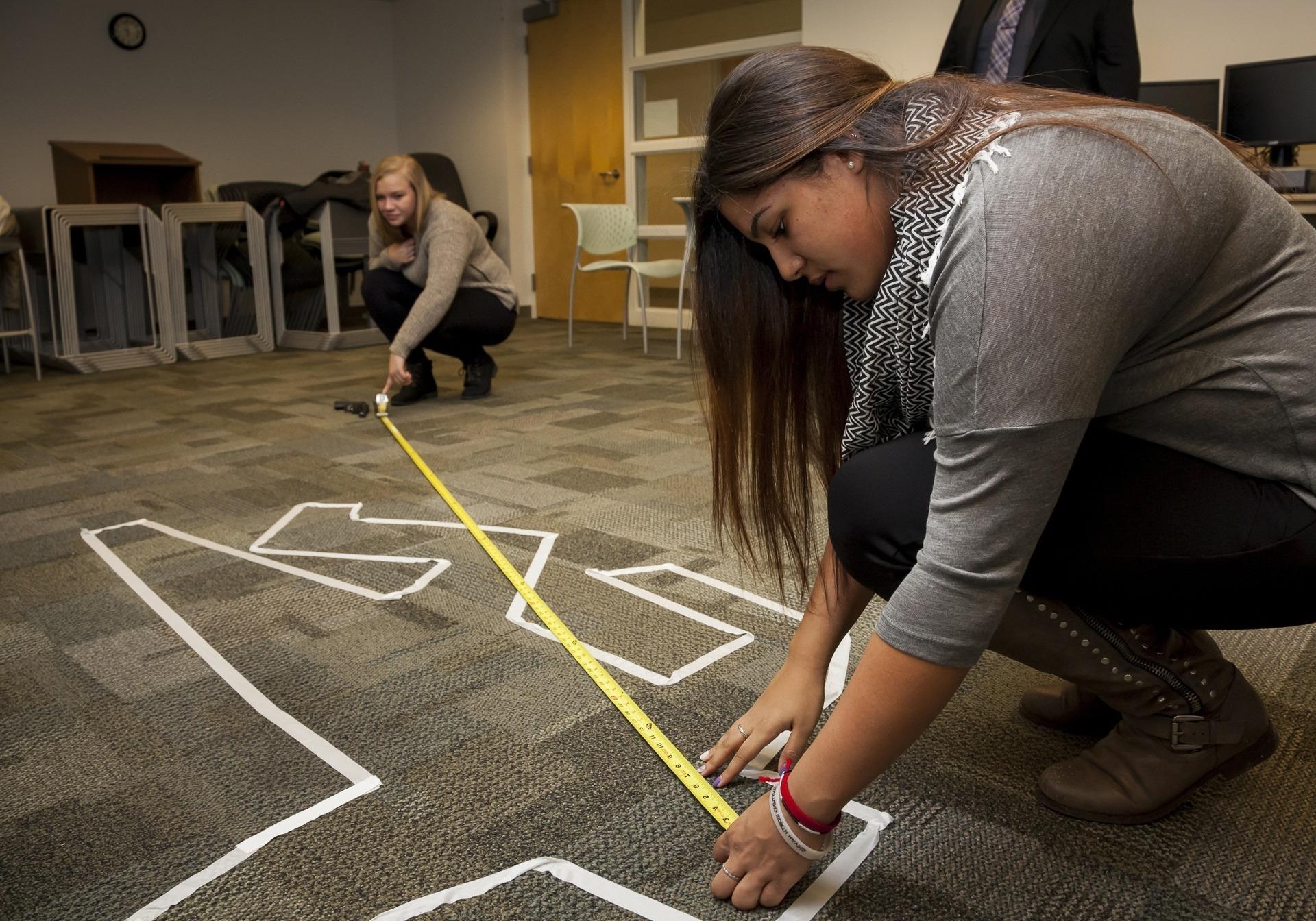 Two female BGSU forensic investigation students measure the distance from a body to a firearm in an on-campus crime scene lab.